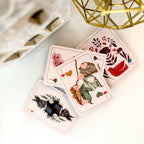 Four illustrated playing cards fanned out on a white surface, with decorative floral and animal artwork and pink borders. A gold geometric object and part of a white box are visible in the background.