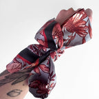 A close-up of a fabric hair scrunchie worn on a person's wrist, featuring a floral pattern in red and pink on a light gray background with black stripes.