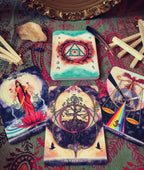 A tarot card deck arranged on a patterned fabric surface with several illustrated cards spread out, including 'Lovers', 'The Poisonous Path', and 'Conjunction'. The deck box shows a green and red design with a triangle and eye symbol.