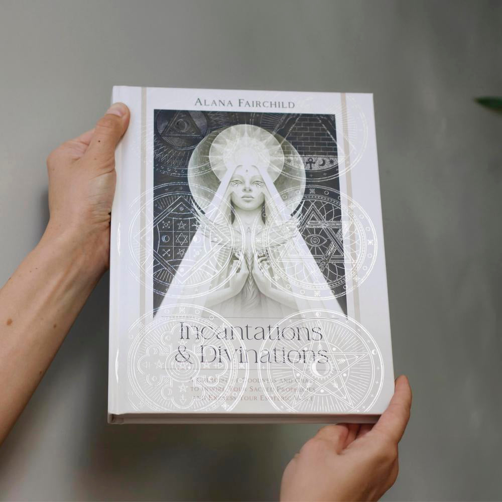 Book titled 'Incentivations & Divinations' by Alana Fairchild held by a person against a neutral background