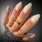 A close-up of a hand with long, glossy almond-shaped nails painted in a warm off-white cream color, wearing gold rings and a gray knit sleeve against a dark gray background.