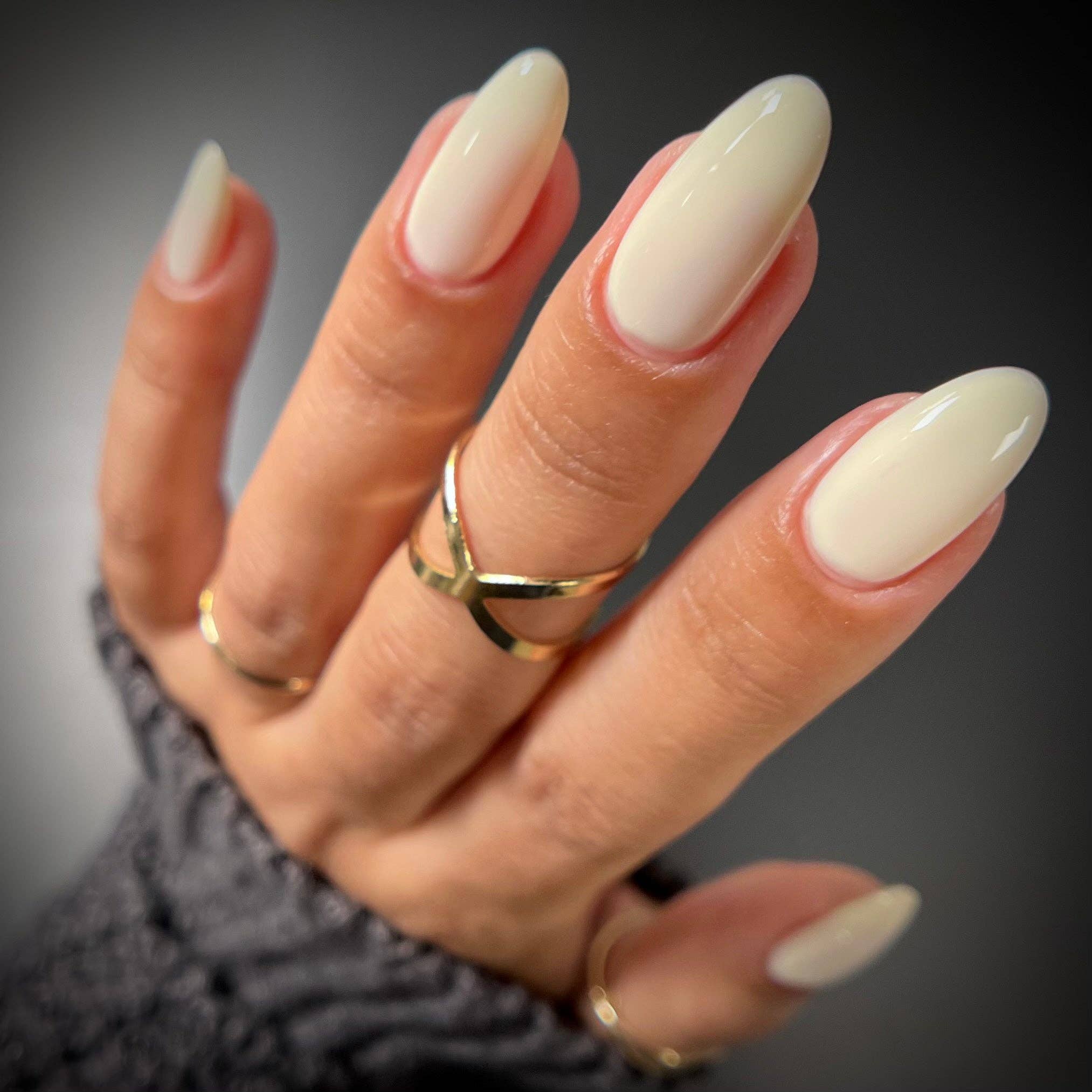 A close-up of a hand with long, glossy almond-shaped nails painted in a warm off-white cream color, wearing gold rings and a gray knit sleeve against a dark gray background.