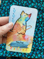 A hand holding a colorful tarot card titled 'Play Time' with a watercolor-style cat illustration, set against a blue patterned fabric background.