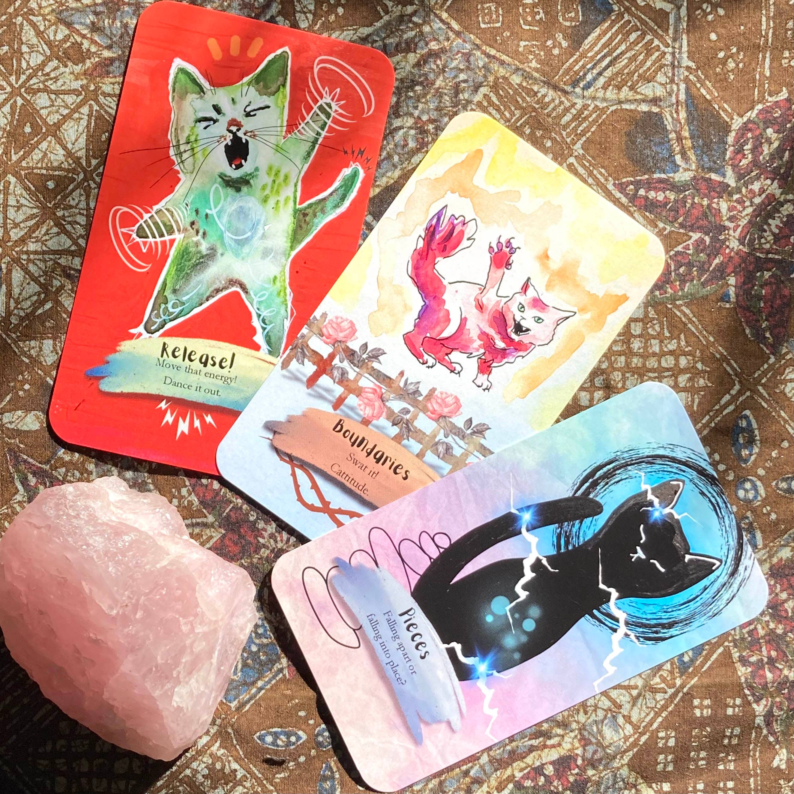 Three illustrated tarot-style cards fanned out on a patterned fabric surface, with a pink crystal rock beside them. The cards feature colorful cat illustrations and text including 'Release!', 'boundaries', and 'pieces'.