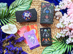 Collection of tarot cards with colorful designs on a woven mat surrounded by plants and flowers.