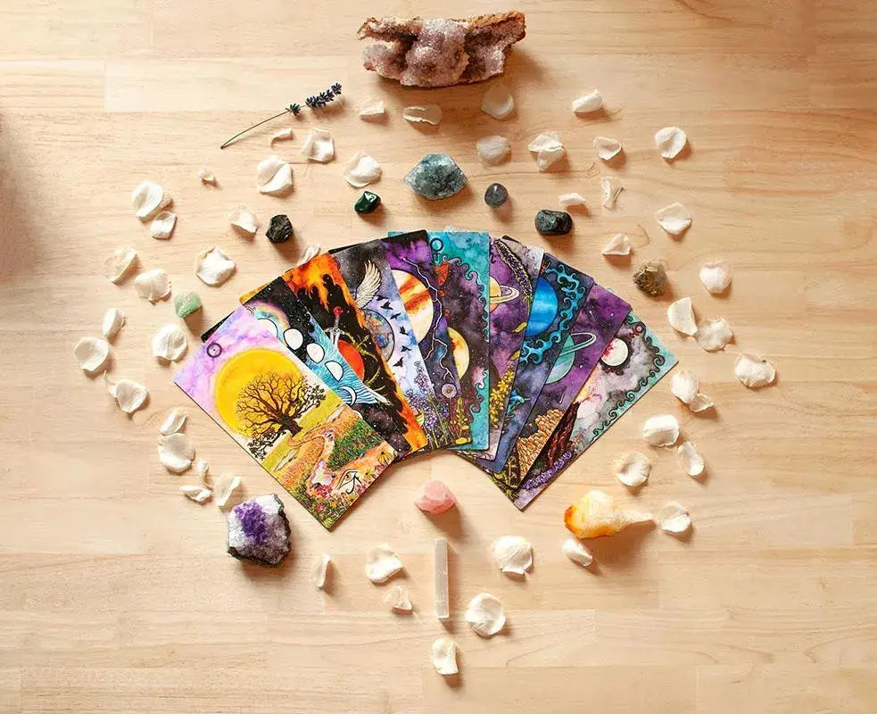 A fan of colorful tarot cards spread out on a light wood surface, surrounded by crystals, stones, and small white petals. The cards feature vibrant, illustrated designs with various colors and symbols.
