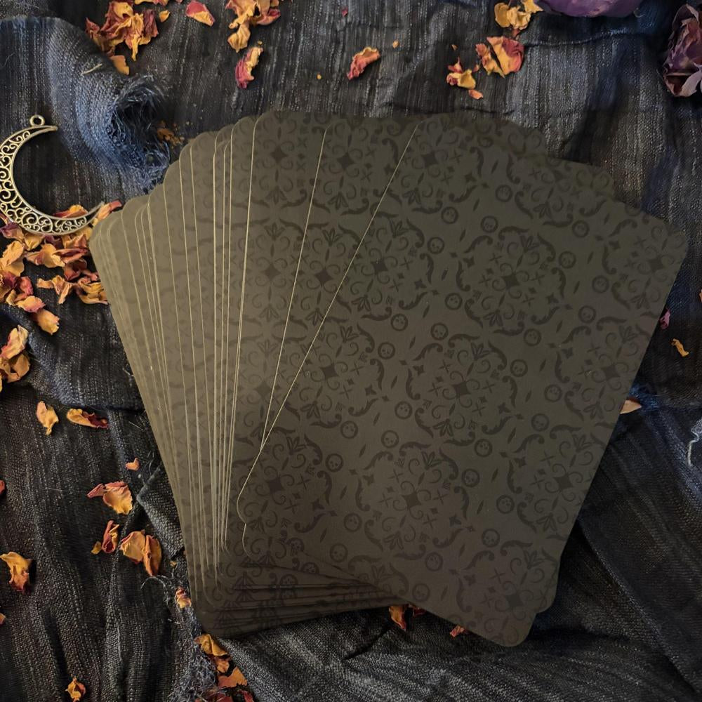 Set of black playing cards with intricate design on a textured surface with dried flowers and a moon symbol.