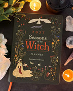 A 2027 'Seasons of the Witch' planner by Lorianne Anderson is displayed in a styled flat-lay setting. The dark green cover features gold and orange illustrations of a witch, a bird, and seasonal motifs.