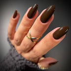 A close-up of a hand with glossy dark brown almond-shaped nails, wearing gold rings and a gray knit sleeve against a dark gray background.