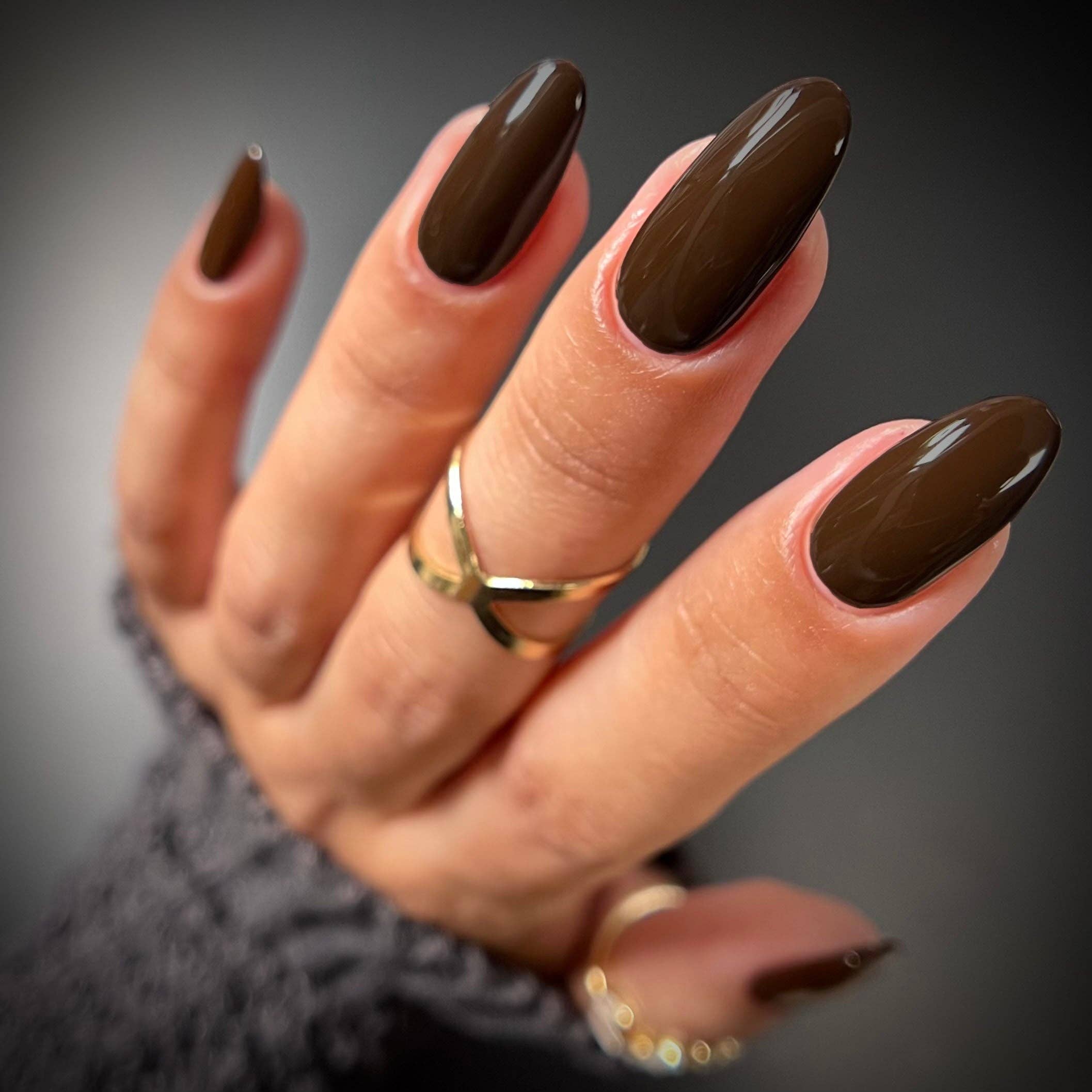 A close-up of a hand with glossy dark brown almond-shaped nails, wearing gold rings and a gray knit sleeve against a dark gray background.