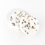 Two round white bath or body soaks with dried botanical pieces embedded, displayed on a clean white background.