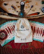 An alchemical card deck set displayed with a beige drawstring pouch printed with 'THE ALCHEMICAL SELE' logo, fanned cards showing teal and red designs, crystals, feathers, and ritual tools arranged on a wooden slice background.