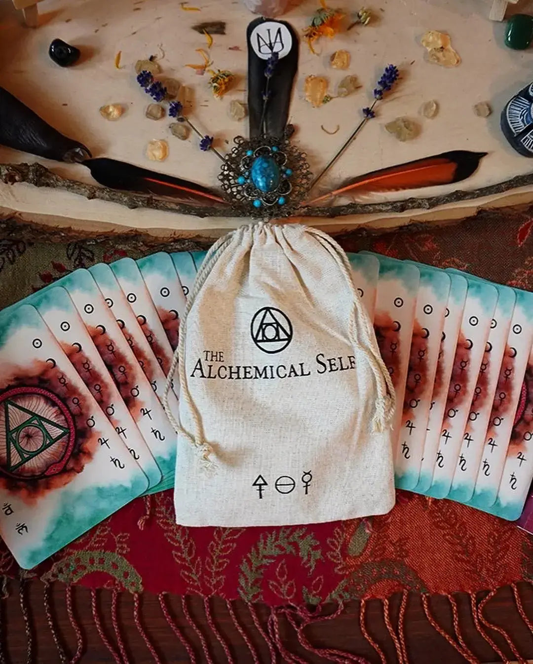 An alchemical card deck set displayed with a beige drawstring pouch printed with 'THE ALCHEMICAL SELE' logo, fanned cards showing teal and red designs, crystals, feathers, and ritual tools arranged on a wooden slice background.