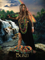 Woman with tattoos and colorful necklaces standing in front of a waterfall with a rainbow, wearing a black dress.