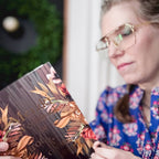 A woman wearing gold-rimmed glasses and a blue floral blouse is holding a book with a dark cover decorated with warm-toned autumn leaves and floral illustrations.