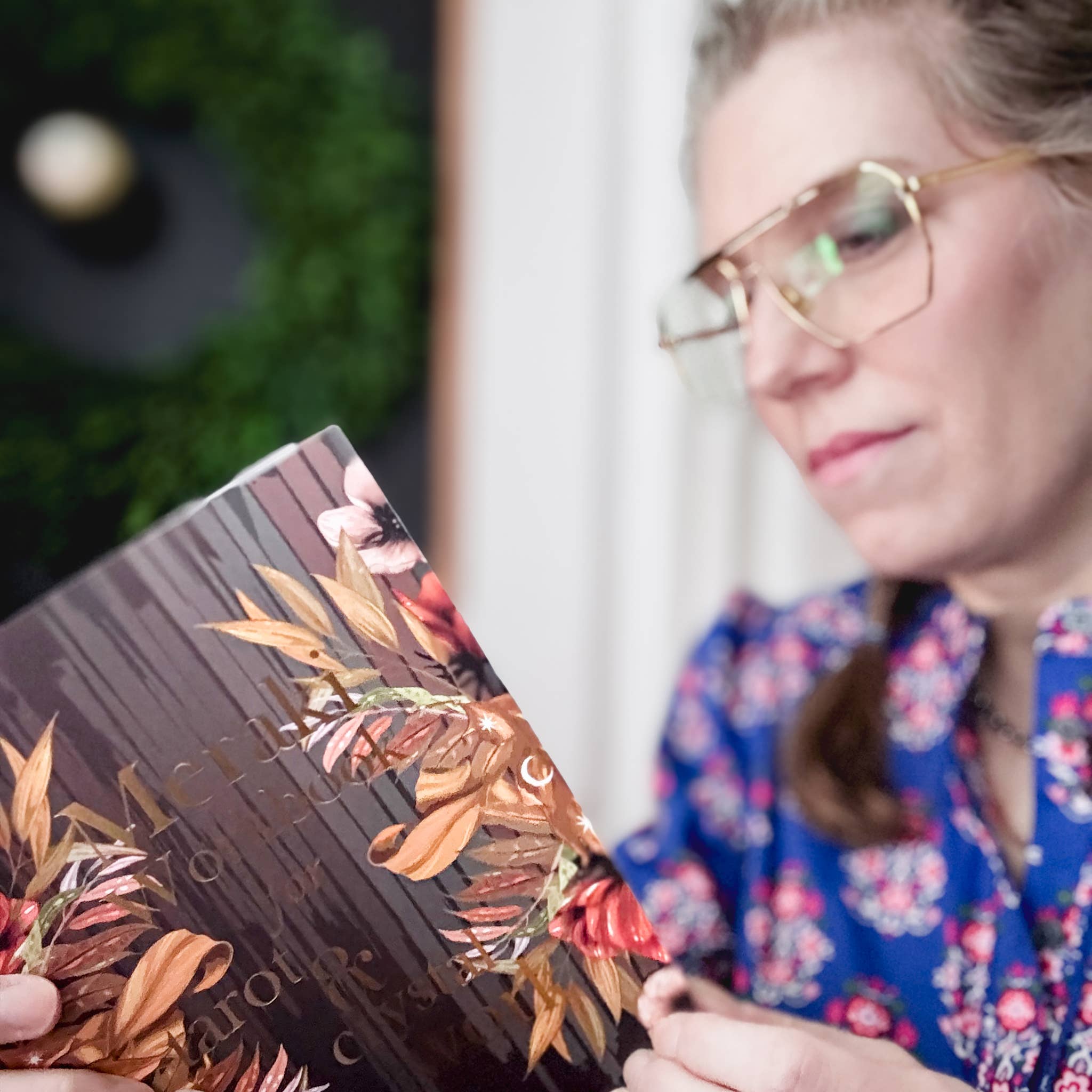 A woman wearing gold-rimmed glasses and a blue floral blouse is holding a book with a dark cover decorated with warm-toned autumn leaves and floral illustrations.