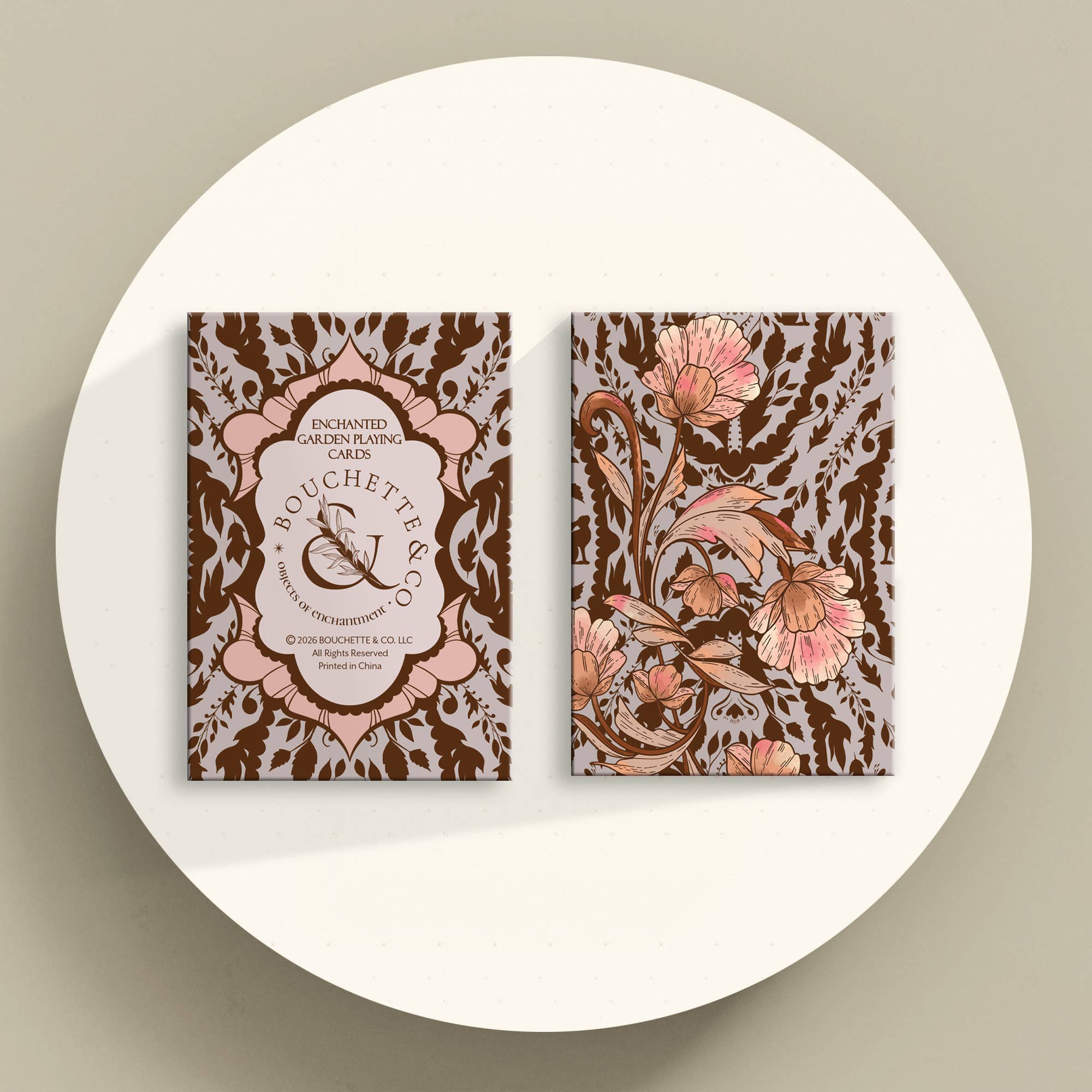 Two decorative greeting or blogging cards displayed upright on a round white surface against a warm beige background. The cards feature ornate floral and damask-style artwork in brown, blush pink, and beige tones, with one card showing a central label reading 'Bouchettes' and the other showing large pink flowers.