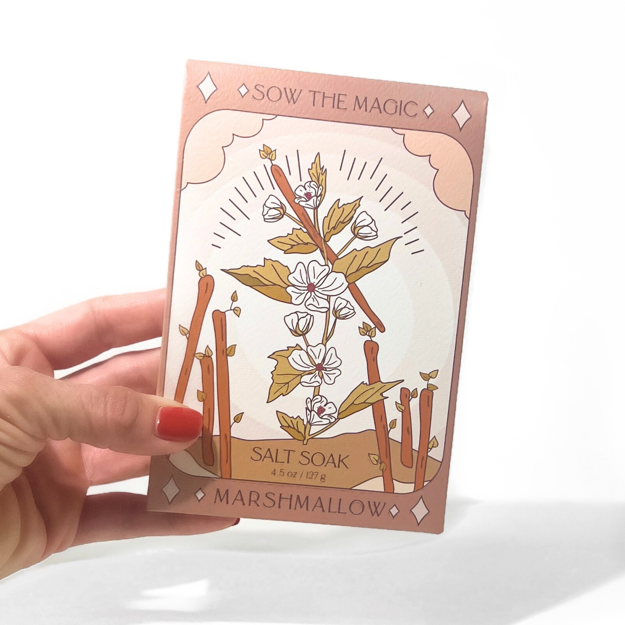 Hand holding a 'Sow the Magic' marshmallow salt soak packaging with floral design.