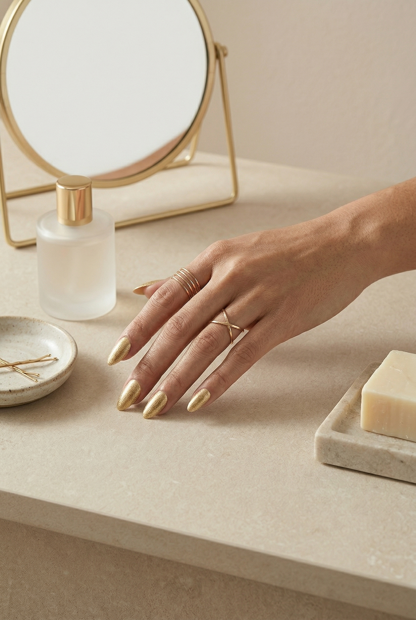 Hand with gold nail polish on a beige surface with a mirror and soap dish.