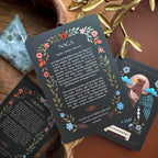 A set of tarot-style oracle cards arranged on a wooden surface, with the central card titled NAGA. The cards have a dark charcoal background with floral borders in pink, orange, blue, and green, and are styled with crystals, a gold branch, and a brown leather-like surface.