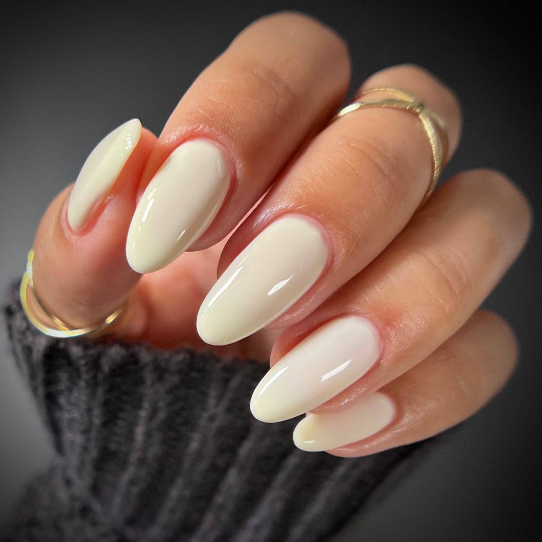 A close-up of a hand with long, glossy almond-shaped nails painted in a warm off-white cream shade. The hand is wearing two thin gold rings and a dark gray ribbed knit sleeve, set against a dark gray background.