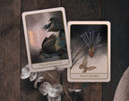 Two illustrated tarot-style cards, titled 'Five of Pentacles' and 'Ten of Swords,' placed on a dark wooden surface with decorative dried leaves and a textured fabric edge nearby.