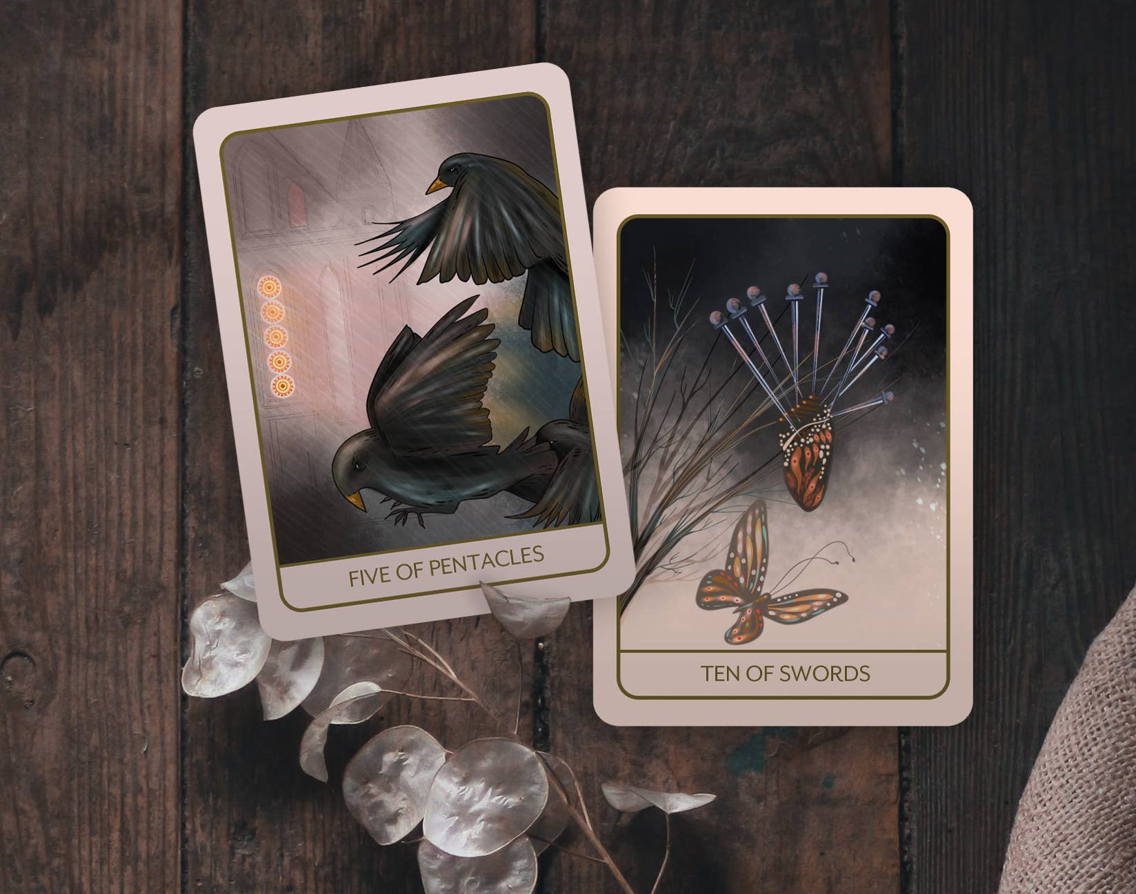 Two illustrated tarot-style cards, titled 'Five of Pentacles' and 'Ten of Swords,' placed on a dark wooden surface with decorative dried leaves and a textured fabric edge nearby.