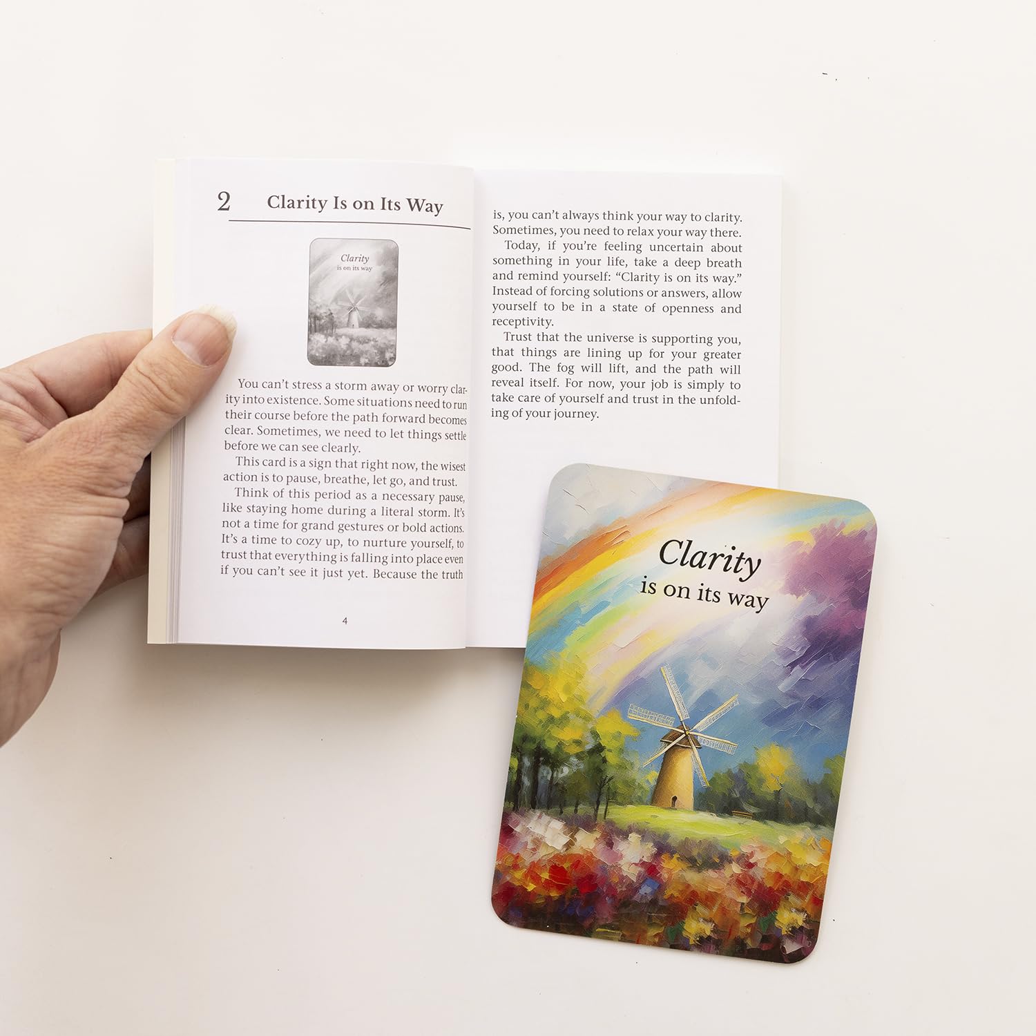 Hand holding an open book with a colorful card featuring a windmill and rainbow titled 'Clarity is on its way' on a white surface.