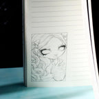 A lined notebook or journal standing upright, showing a black-and-white pencil sketch of a stylized girl with closed eyes on one page, photographed against a dark background with a light blue surface below.
