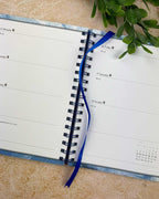 An open spiral-bound planner or agenda with white pages, a blue ribbon bookmark, and a blue patterned cover edge, photographed on a beige stone-like surface with green leaves at the top.
