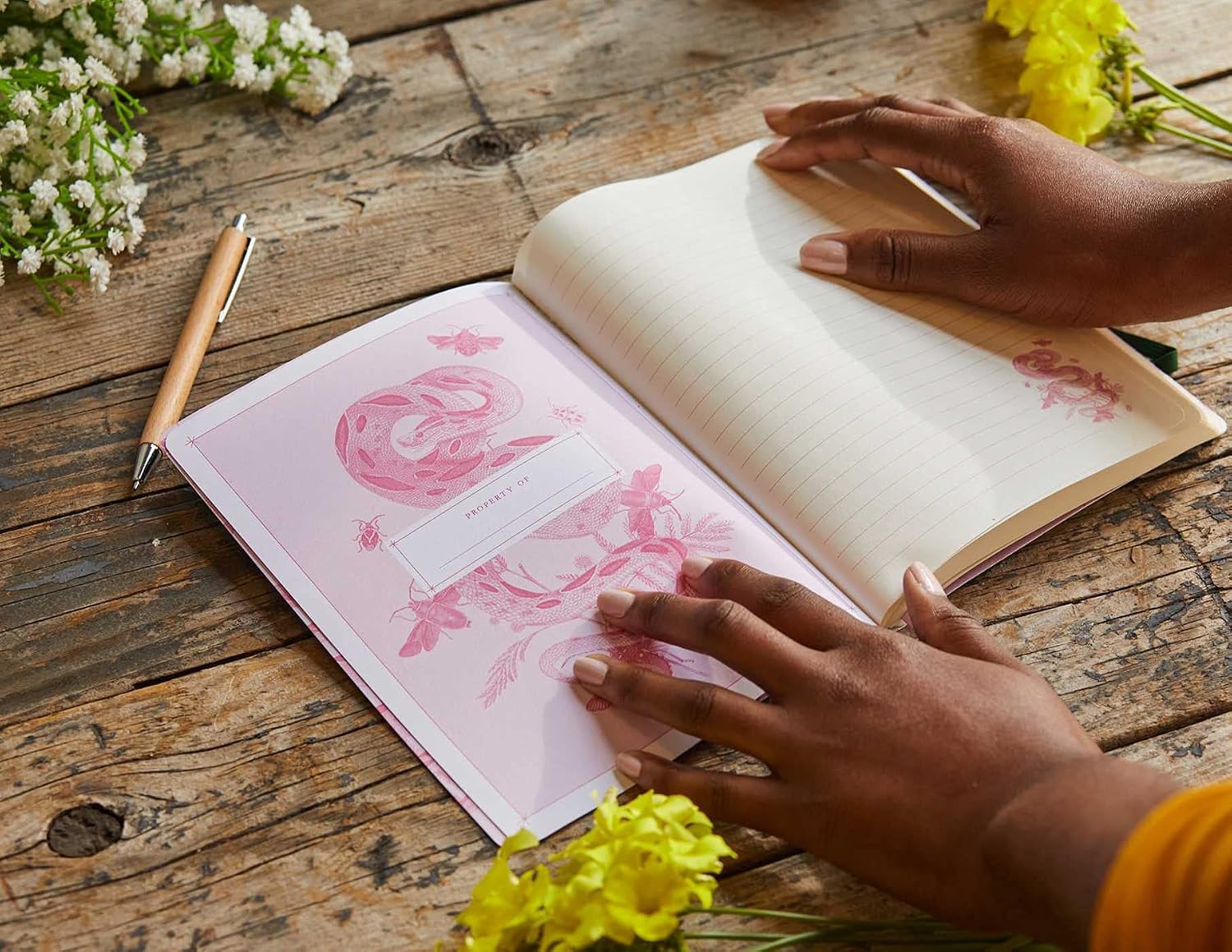 An open journal or notebook with pink floral artwork on the left page, resting on a rustic wooden table. Two hands are visible holding the open book, with small white and yellow flowers placed around it.