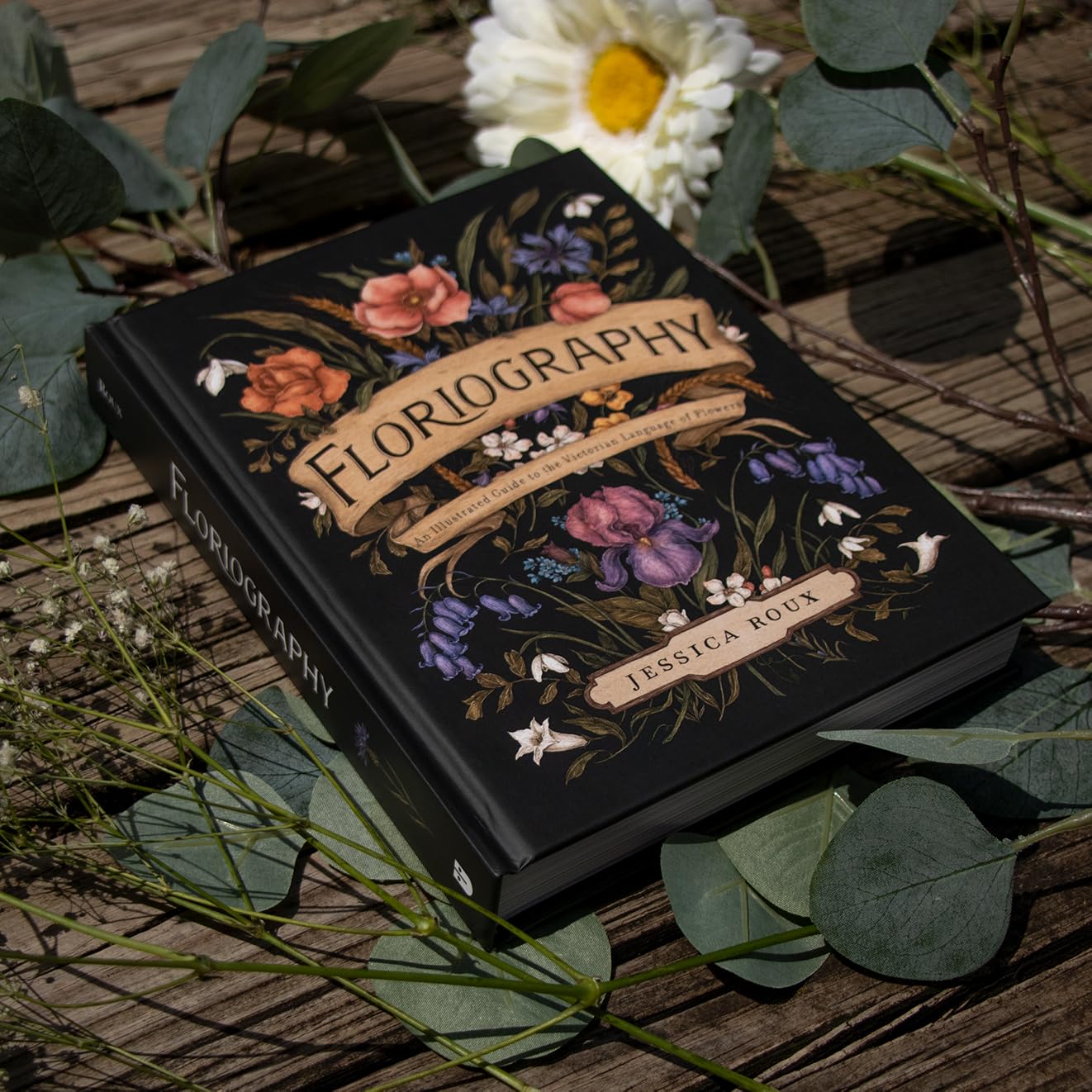 Book titled 'Floriography' by Jessica Roux with floral designs on a wooden surface with leaves and flowers.