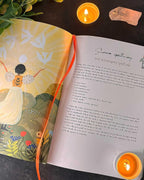 An open illustrated book with a printed spellbook page titled 'Summer apothecary Self-healing spell ink' spread open, styled with two lit orange candles, a pink crystal, and green leaves on a dark surface.