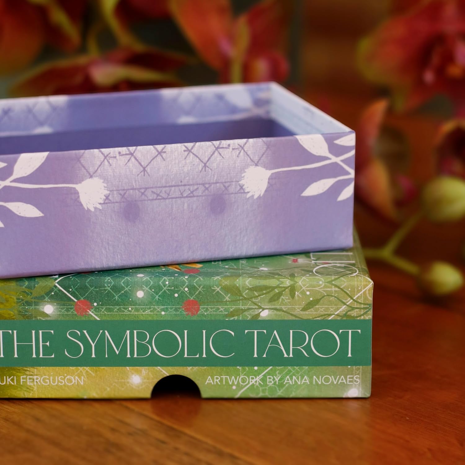 Two tarot card boxes, one purple and one green with text, on a wooden surface with flowers in the background.