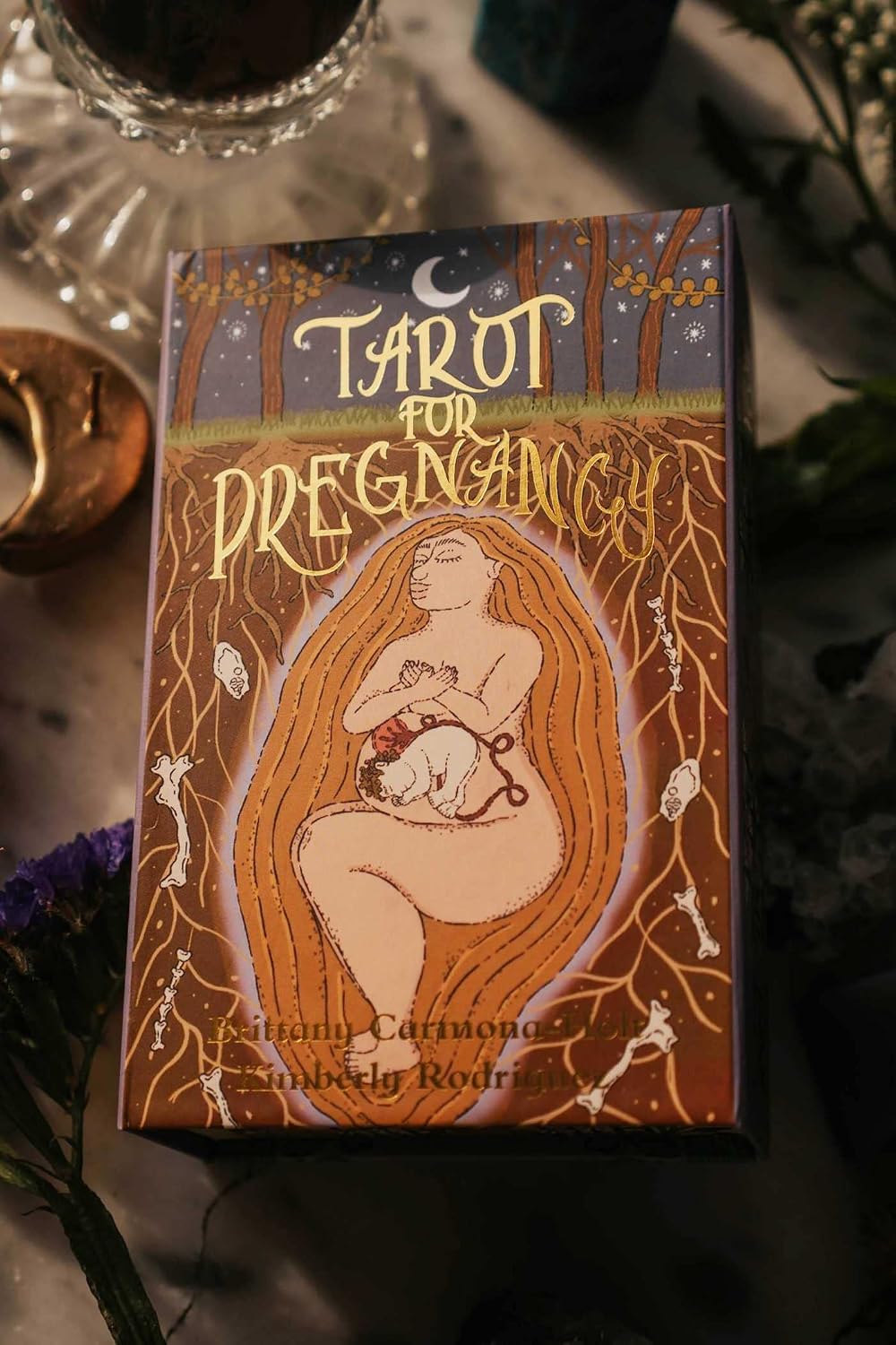 A boxed tarot card deck titled 'Tarot for Pregnancy' sits on a tabletop. The box features an illustrated nude woman with long flowing hair holding a baby, surrounded by roots and stars, with the text 'Many Cycles & Many Beginnings' at the bottom.