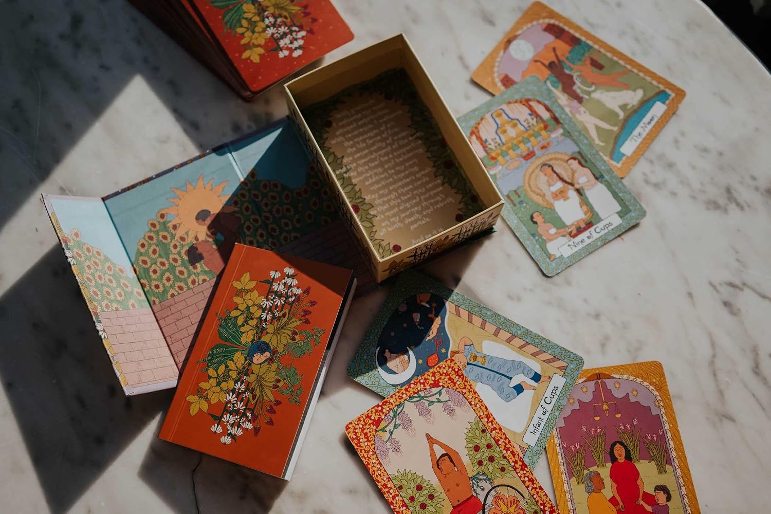 A set of illustrated tarot cards and an open decorative box arranged on a light marble surface in natural sunlight.