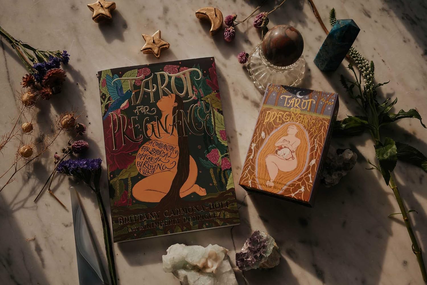 Two tarot deck boxes for pregnancy-themed card games, styled in a flat lay with crystals, dried flowers, and wooden stars on a marble surface.