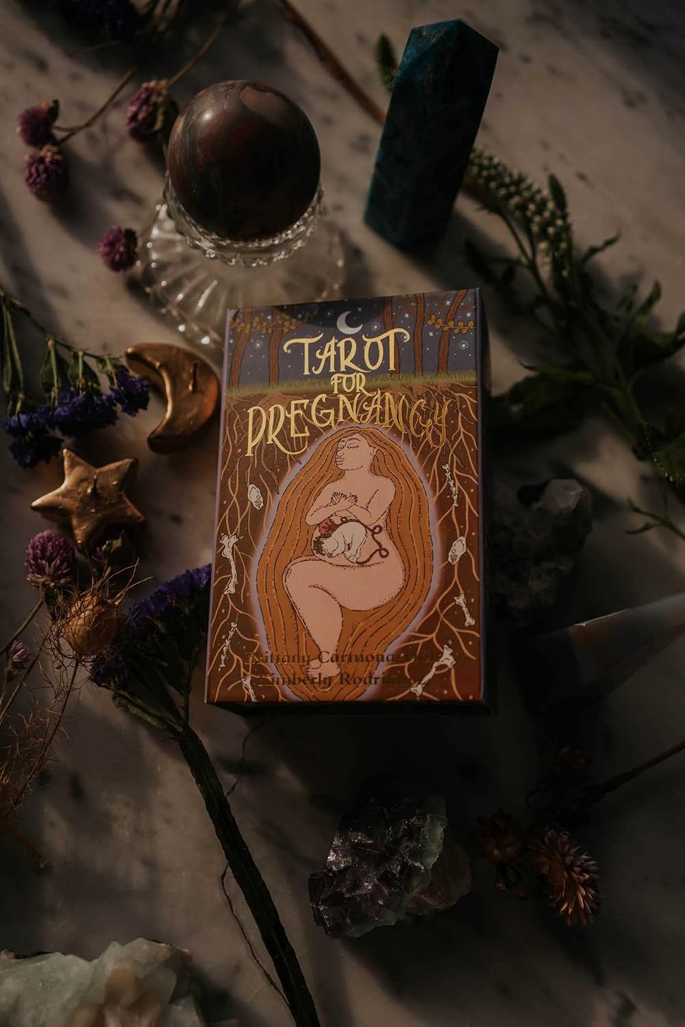 A boxed tarot card deck titled 'Tarot for Pregnancy' is displayed on a styled surface with crystals, dried flowers, a glass sphere, and candles. The box features an illustration of a pregnant woman holding a baby, with warm orange, brown, and cream tones.