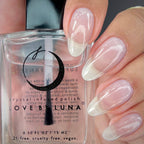 A close-up of a hand with glossy almond-shaped nails painted in a soft pale pink and white ombre style, holding a bottle of Love by Luna Crystal Infused Polish.