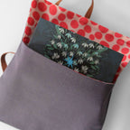 A fabric tote bag shown open, with a dark gray front pocket holding an illustrated card featuring white flowers and a blue star. The bag has red polka dots on a light background on the inside and brown handles, photographed on a plain light background.