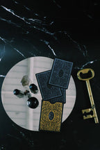 A styled flat-lay featuring a deck of black playing cards with ornate gold patterns, three fanned cards, a large antique gold key, and a small cluster of crystals on a white circular mat over a black marble surface.