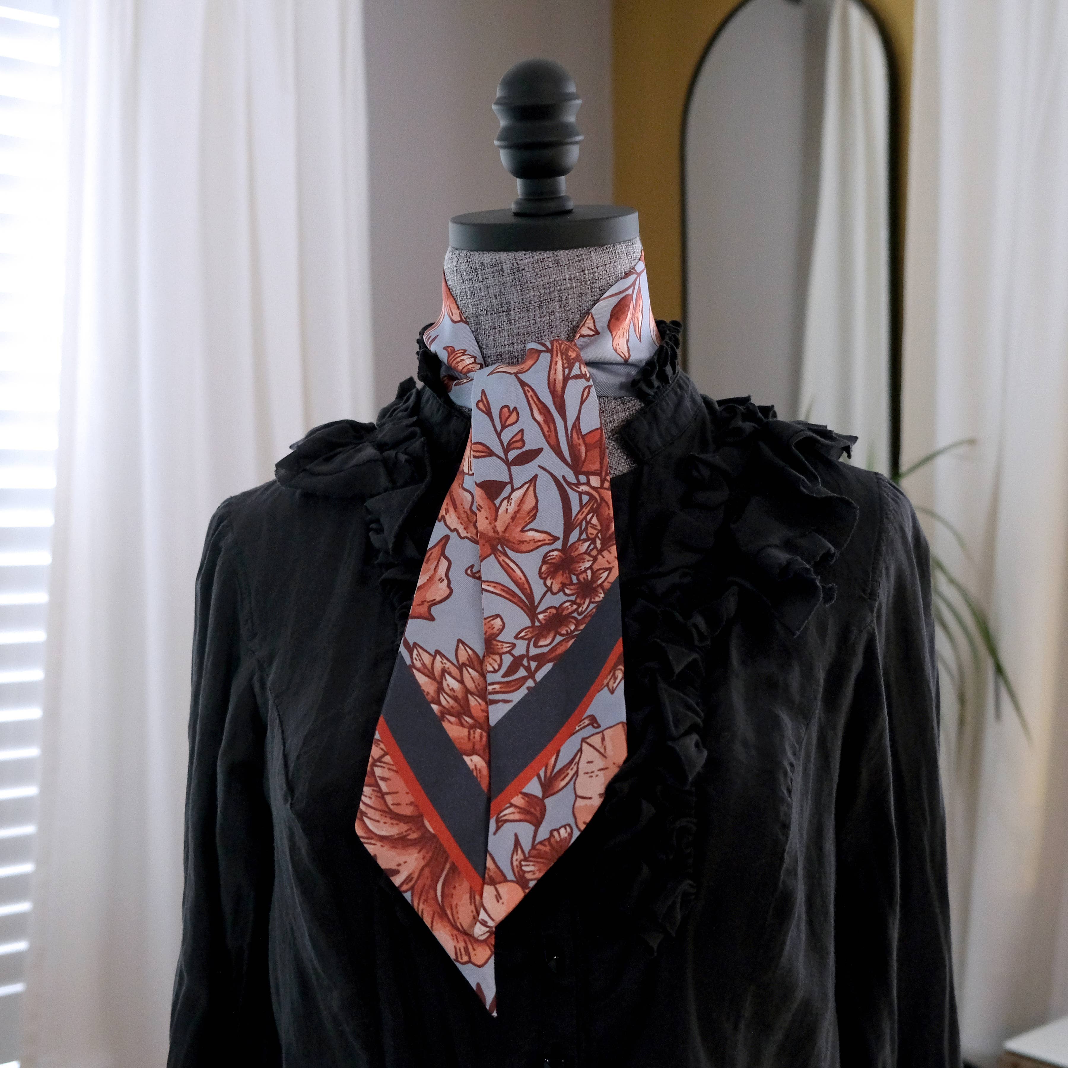Mannequin wearing a black outfit with a floral patterned scarf in a room with white curtains.