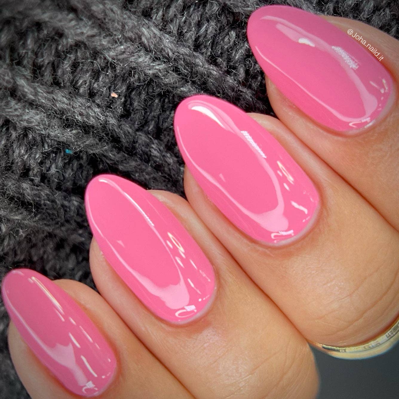 A close-up of a hand with long, glossy pink manicured nails resting against a dark gray knitted background.