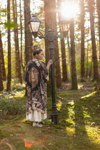 Woman in a patterned dress standing next to a lamp post in a forest with sunlight filtering through the trees.