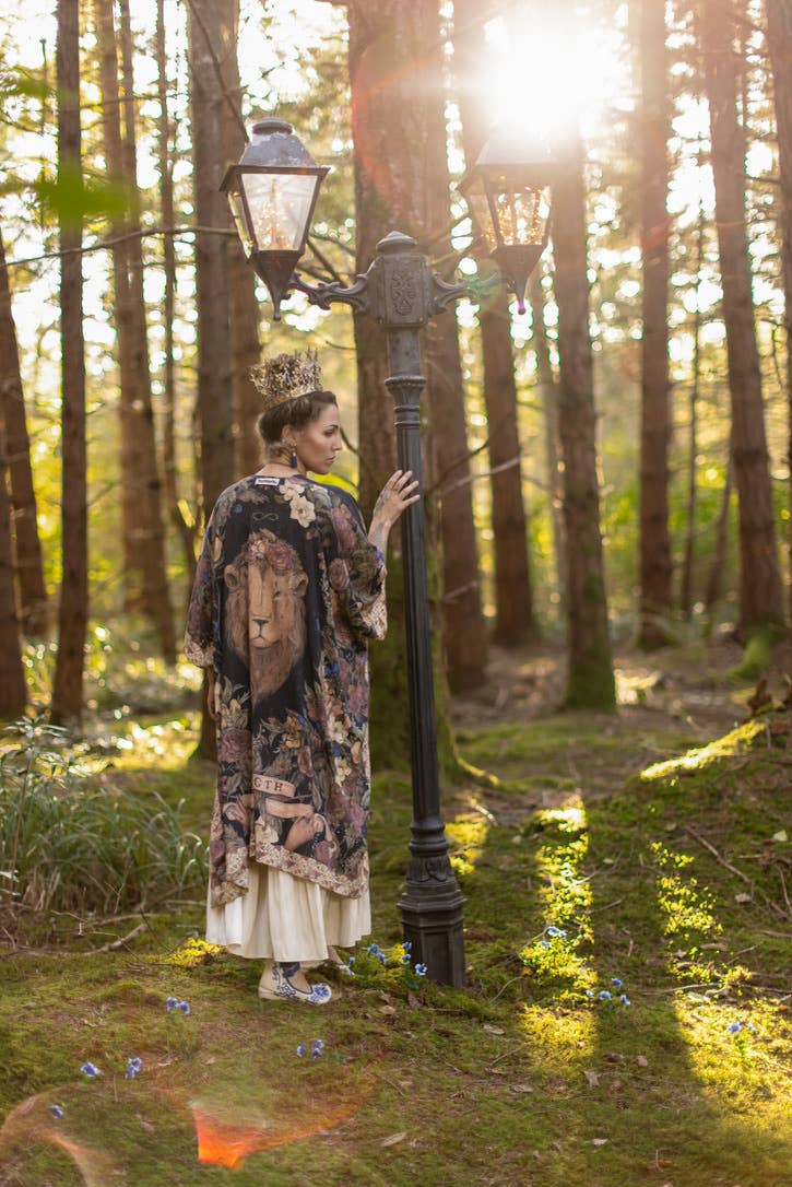 Woman in a patterned dress standing next to a lamp post in a forest with sunlight filtering through the trees.