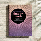 The book cover of 'Shadow Work' by Danielle Massi, LMFPT, featuring a black central circle with pink text, surrounded by a pink-to-purple sunburst line pattern. The book is photographed standing upright on a textured, light gray stone surface with soft shadows.