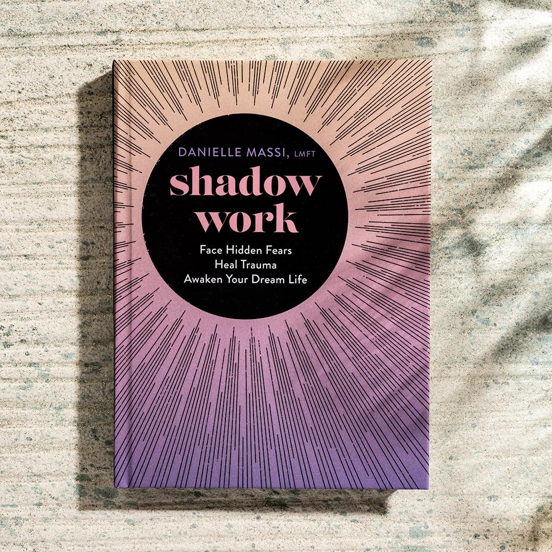 The book cover of 'Shadow Work' by Danielle Massi, LMFPT, featuring a black central circle with pink text, surrounded by a pink-to-purple sunburst line pattern. The book is photographed standing upright on a textured, light gray stone surface with soft shadows.