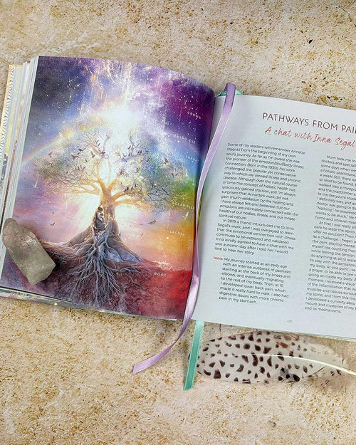 An open book or journal lying on a textured beige surface, showing a colorful tree-of-life illustration on one page and printed text on the other. A purple ribbon bookmark hangs from the right page, and a small gray stone and a feather are placed beside it.