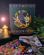 Kate Forsyth's Hedgewitch Oracle card deck box standing upright on a dark surface, surrounded by illustrated cards, a lit candle, and green foliage. The box features a dark green background with a central illustration of a black raven perched on a branch under a crescent moon.