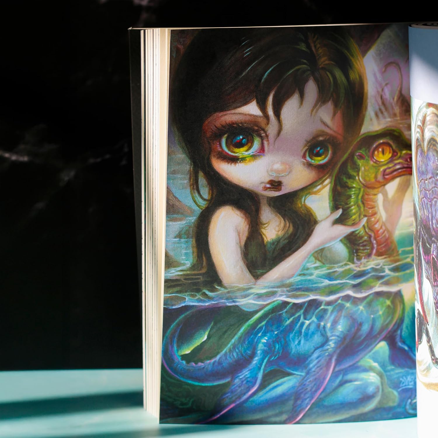A close-up of a colorful illustrated book or art print showing a fantasy mermaid-like girl with large green eyes, dark hair, and a blue-green fish or sea creature, photographed against a dark background.